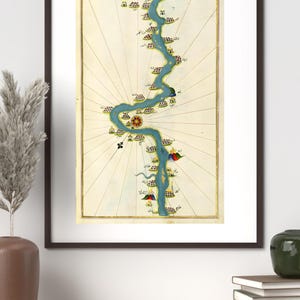 Puede incluir: Impresión enmarcada de un mapa antiguo que representa un río azul sinuoso con ilustraciones detalladas de edificios y puntos de referencia. El mapa está sobre un fondo crema con líneas radiales, todo dentro de un marco marrón oscuro.