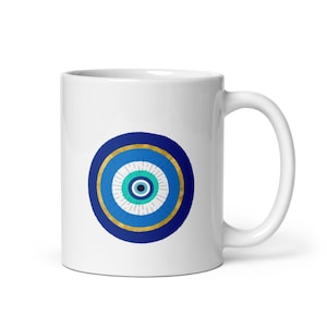 May include: White ceramic mug featuring a blue and gold evil eye design. The eye symbol has concentric circles of blue, turquoise, and white, with a black pupil. The mug has a curved handle.