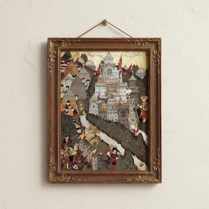 May include: Framed artwork depicting a detailed historical scene with a fortress, figures in period clothing, and red flags. The frame is ornate, brown, and rectangular, with a gold-colored accent. The artwork is mounted on a cream-colored wall.