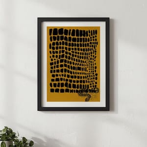 Puede incluir: Impresión artística enmarcada con un diseño en negro y dorado. La obra de arte muestra un patrón texturizado que se asemeja a la piel de reptil, con una ilustración detallada de una serpiente en la parte inferior. La impresión está en un marco negro, ideal para la decoración de paredes.