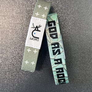 May include: Two woven wristbands. One is green with white plus signs and a logo that says "EWOPT". The other is blue and green with a white floral pattern and the text "GOOD AS A ROGUE".