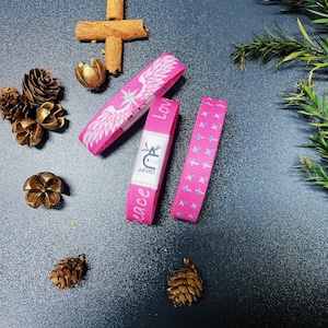 May include: Three pink fabric wristbands with different designs. One has a white angel wing design with the word "Love" on it. Another has a blue and pink cross design. The third has the word "Peace" and the word "Effort" on it.
