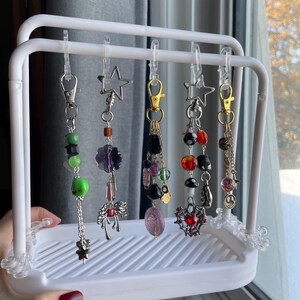 May include: A white metal display rack holding several decorative keychains. The keychains feature various charms, including stars, beads, and pendants in colors like green, purple, black, and red. The rack is held by a hand with red nail polish.