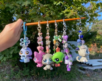May include: A collection of colorful keychains featuring various animal figurines. Each keychain has a unique design, with charms and beads in shades of pink, green, purple, and blue. The keychains are suspended from a wooden dowel.