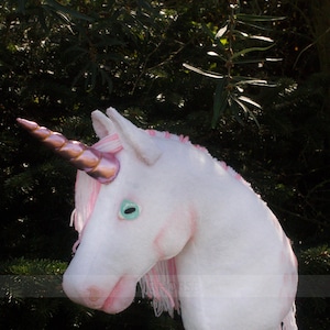 Peut inclure: Une tête de licorne blanche avec une crinière rose et une corne dorée en spirale. La licorne a un œil bleu clair et des joues roses. La tête est fixée à un bâton en bois.