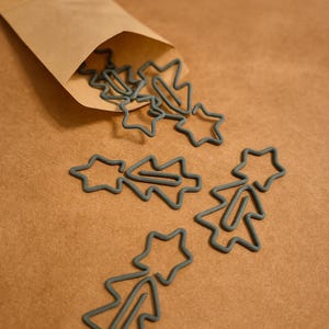 Könnte beinhalten: Graue Metall-Büroklammern in Form von Weihnachtsbäumen und Sternen, die aus einer braunen Papiertüte fallen. Die Büroklammern sind auf einer braunen Oberfläche verstreut.