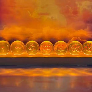 以下が含まれることがあります： 赤色の星が入った、7つの光る半透明の球体が木製の台座に配置されています。 球体は内側から照らされ、暖かく琥珀色の輝きを生み出しています。 背景にはオレンジと黄色の空が広がっています。