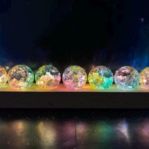 May include: Seven spherical glass orbs with a cracked, iridescent texture arranged in a horizontal row on a rectangular wooden base. Each orb glows with internal multicoloured light, displaying hues of pink, blue, green, and yellow.