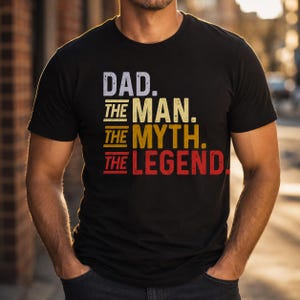 以下が含まれることがあります： 「DAD. THE MAN. THE MYTH. THE LEGEND」の文字が白、黄色、赤でプリントされた黒いTシャツ。クルーネックの半袖Tシャツです。