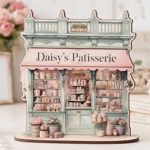 May include: A wooden model of "Daisy's Patisserie", featuring a pink awning and shelves stocked with miniature pastries. The building is painted in pastel shades, with flower boxes and decorative details. The overall design is charming.