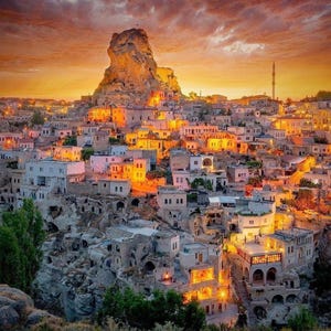 May include: A scenic view of a town nestled in a valley, illuminated by warm lights against a sunset sky. The town features white buildings with orange and yellow lights, and a large rock formation in the background.