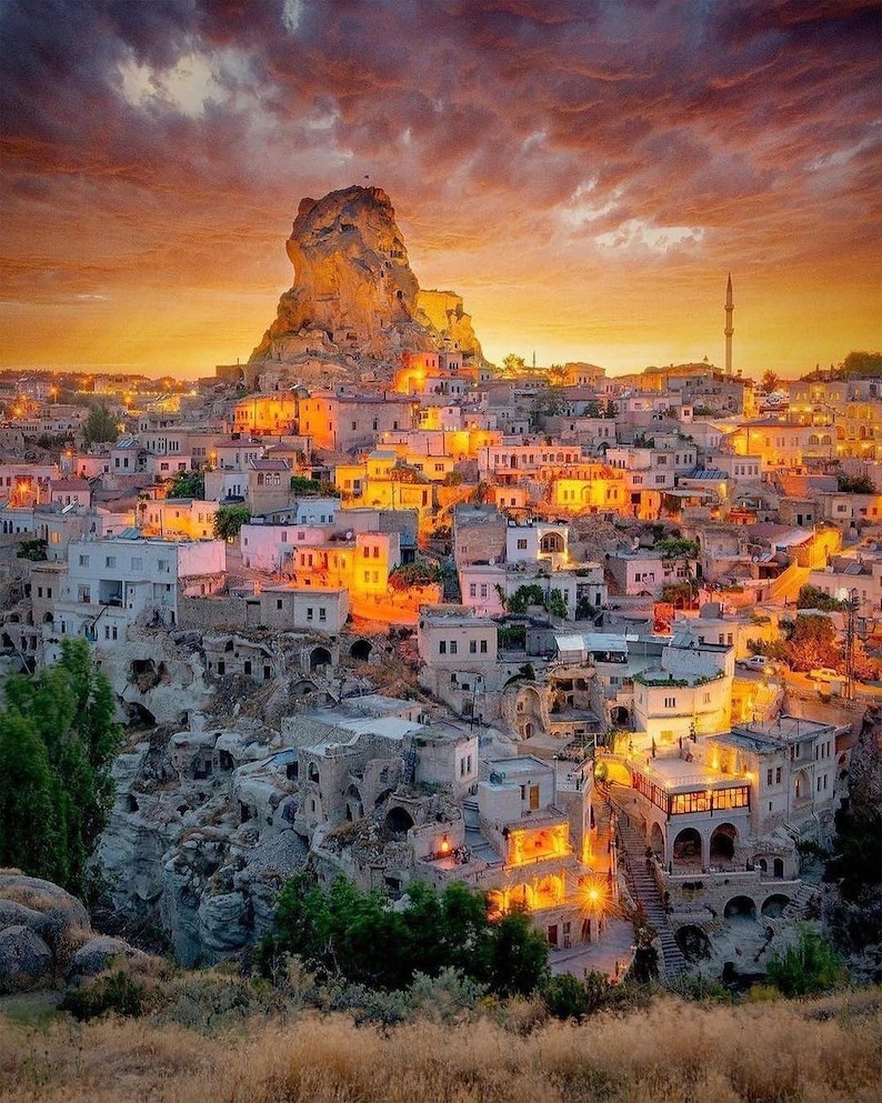 May include: A scenic view of a town nestled in a valley, illuminated by warm lights against a sunset sky. The town features white buildings with orange and yellow lights, and a large rock formation in the background.