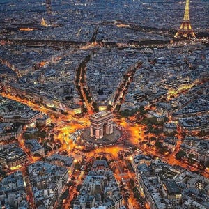 May include: Aerial view of Paris at dusk, showcasing the illuminated Arc de Triomphe at the center, surrounded by radiating avenues and city lights. The Eiffel Tower is visible in the distance, with a dark blue sky.