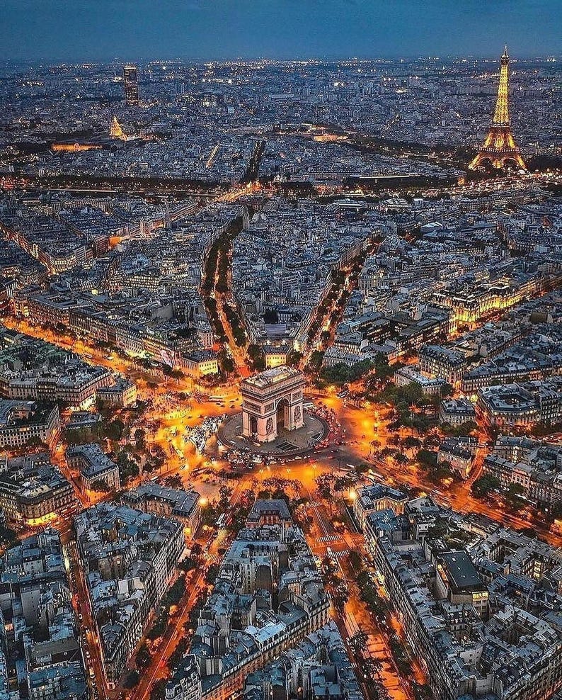 May include: Aerial view of Paris at dusk, showcasing the illuminated Arc de Triomphe at the center, surrounded by radiating avenues and city lights. The Eiffel Tower is visible in the distance, with a dark blue sky.