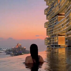 May include: A person in a pool at sunset, with a city skyline in the distance. A modern building with illuminated windows is on the right. The sky is a gradient of orange and pink.