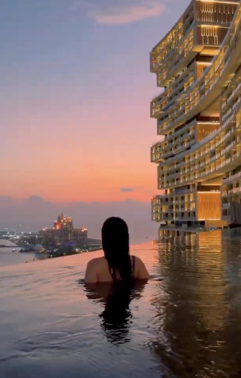 May include: A person in a pool at sunset, with a city skyline in the distance. A modern building with illuminated windows is on the right. The sky is a gradient of orange and pink.