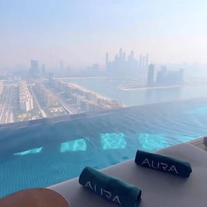 May include: An aerial view of a rooftop pool with a cityscape in the background. Two rolled-up teal towels with the word "AURA" are on a lounge chair. A round, brown table is in the foreground.