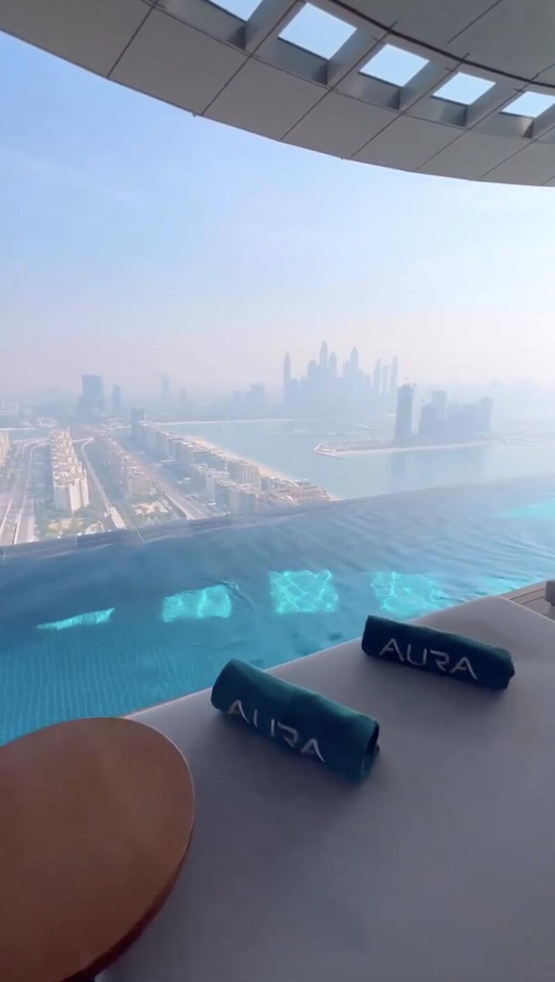 May include: An aerial view of a rooftop pool with a cityscape in the background. Two rolled-up teal towels with the word "AURA" are on a lounge chair. A round, brown table is in the foreground.