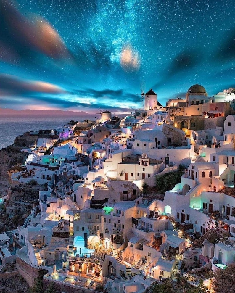 May include: A picturesque nighttime view of a coastal village with white buildings and windmills under a starry sky. The buildings are illuminated with warm lights, creating a contrast with the cool blues and purples of the sky and sea.