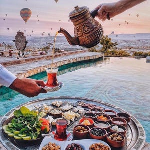 May include: A scenic outdoor breakfast setting with a large silver tray filled with various foods, including cheeses, olives, and nuts. A person pours tea from a decorative copper teapot into a glass. Hot air balloons float in the background.