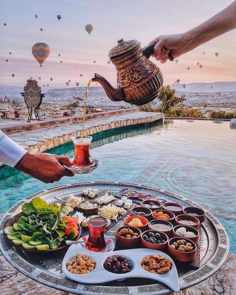 May include: A scenic outdoor breakfast setting with a large silver tray filled with various foods, including cheeses, olives, and nuts. A person pours tea from a decorative copper teapot into a glass. Hot air balloons float in the background.