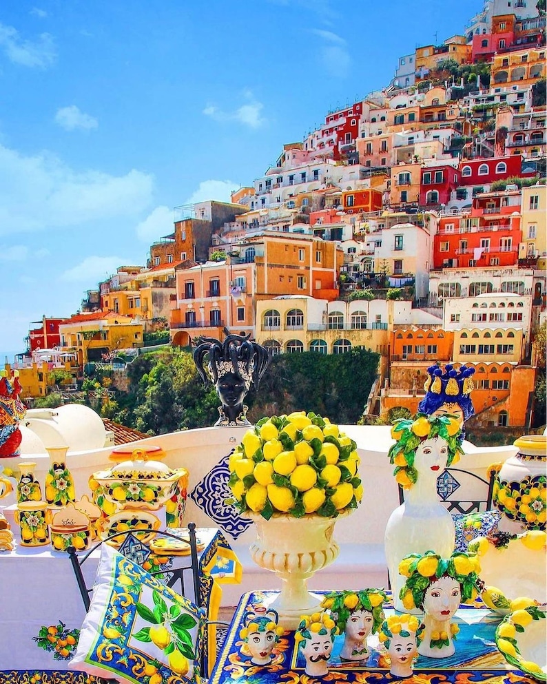 May include: A vibrant display of ceramic art and decorative items, including vases, bowls, and head sculptures, with a lemon theme. The background features a colorful hillside town under a clear blue sky.