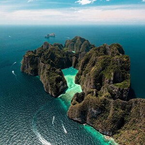 May include: Aerial view of a tropical island with lush green vegetation and rocky cliffs surrounded by turquoise and deep blue water. Several boats are visible in the water, and the sky is a clear blue with a few clouds.