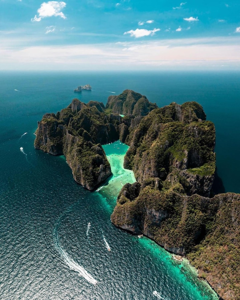May include: Aerial view of a tropical island with lush green vegetation and rocky cliffs surrounded by turquoise and deep blue water. Several boats are visible in the water, and the sky is a clear blue with a few clouds.