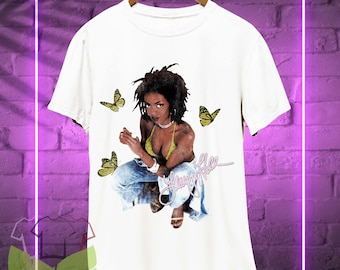 May include: White t-shirt featuring a graphic of a woman in a yellow bikini top and blue jeans, with yellow butterflies. The text "Lauryn Hill" is written in a cursive font. The shirt is hanging on a wooden hanger.