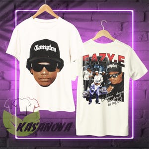 Puede incluir: Dos camisetas blancas colgadas en perchas de madera. Una presenta un retrato de un hombre con gorra de Compton y gafas de sol. La otra muestra una foto de Eazy-E con el texto "EAZY-E" y "Cruisin' down the street in my 64".