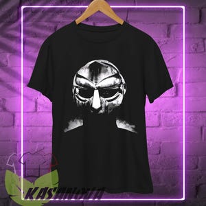 Könnte beinhalten: Schwarzes T-Shirt mit einem Graustufenbild einer Maske. Die Maske hat ein metallisches Aussehen mit markanten Augen- und Munddetails. Das T-Shirt hängt an einem Holzbügel vor einer lila Backsteinmauer.