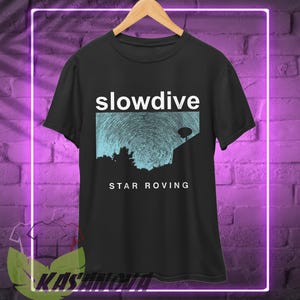 Puede incluir: Camiseta negra con el nombre de la banda "slowdive" en letras blancas sobre un gráfico azul y blanco de un cielo nocturno. El texto "STAR ROVING" está debajo. La camiseta está colgada en una percha de madera.