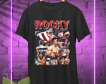 May include: Black t-shirt featuring a collage of images from the movie "Rocky," with the title in red block letters. The design includes action shots of the main character in boxing scenes, with red and white boxing gloves.