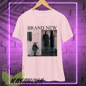 Peut inclure: T-shirt blanc avec l'inscription "BRAND NEW" au-dessus d'une photographie en noir et blanc de trois personnes. La photo présente une scène vintage de personnes devant une maison.