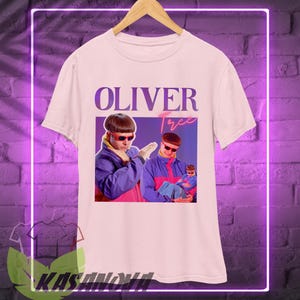 May include: White t-shirt featuring a graphic of Oliver Tree in a purple and blue jacket and red sunglasses. The text "OLIVER Tree" is printed above the image in purple and pink.