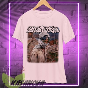 May include: A light pink t-shirt featuring a graphic of a person wearing goggles and a head covering, with the word "AYATOPIA" in stylized text above the image. The shirt is hanging on a wooden hanger.
