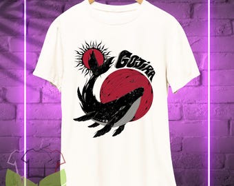 May include: White t-shirt featuring a black and red graphic of a whale breaching in front of a red sun. The word "GOJIRA" is printed above the whale in black, stylized lettering. The shirt is hanging on a wooden hanger.