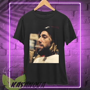 May include: Black t-shirt with a portrait of a person with tattoos, a nose ring, and a cigarette. The person is wearing a black beanie and a white shirt. The image is set against a purple brick wall.