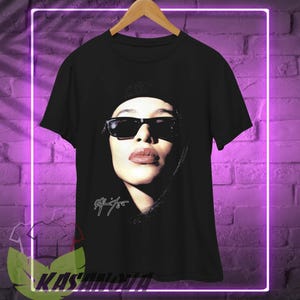 May include: Black t-shirt with a portrait of a woman wearing sunglasses. The image is in black and white with a white signature. The t-shirt is displayed on a wooden hanger.