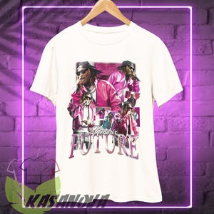 May include: Cream-colored t-shirt featuring a graphic of a person wearing a pink jacket and sunglasses, with the text "Hendrix FUTURE" in a vintage style. The shirt is hanging on a wooden hanger.