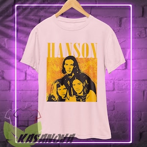 May include: White t-shirt featuring the band Hanson with the band's name in yellow block letters above a yellow and orange image of the band members. The shirt is hanging on a wooden hanger.