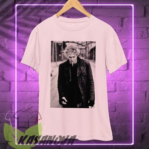 May include: White t-shirt featuring a black and white image of a person in a leather jacket smoking a cigarette. The image is centered on the shirt. The shirt is hanging on a wooden hanger against a purple brick wall. The word "KASANOVA" is at the bottom.