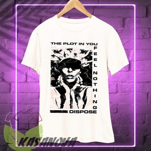 May include: White t-shirt with a graphic design featuring a black and white portrait and the text "THE PLOT IN YOU FEEL NOTHING DISPOSE". The shirt is hanging on a wooden hanger.
