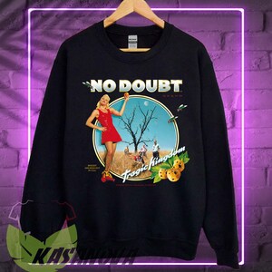 May include: Black sweatshirt featuring a graphic from the band No Doubt's album, Tragic Kingdom. The design includes a woman in a red dress, a tree, and the album title. The sweatshirt is hanging on a wooden hanger.
