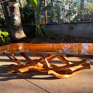 May include: A handcrafted wooden coffee table with a natural, organic design. The tabletop is a single piece of polished wood, supported by a base of intertwined branches. The table's warm, natural tones and unique shape make it a striking centerpiece.