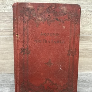 May include: A vintage book titled "AROUND THE TEA TABLE" with a distressed red cover. The cover features ornate black decorative elements and text. The book's spine is visible, and the overall aesthetic suggests an antique or historical item.