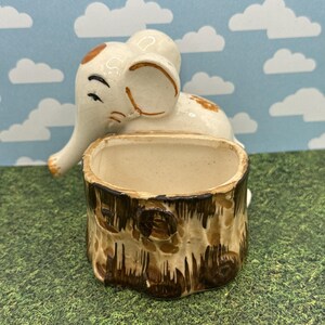 May include: A ceramic elephant planter with a trunk and ears, in cream and brown tones. The planter has a tree trunk design. The elephant is perched above the planter, adding a whimsical touch. The planter is suitable for small plants or decorative items.