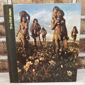 May include: A hardcover book titled "The First Men" with a cover illustration depicting early humans in a grassy field. The image shows several figures, some carrying items, under a blue sky with clouds. The book's spine is dark green.