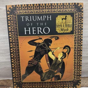 May include: A hardcover book titled "TRIUMPH OF THE HERO" with a black and gold cover. The cover features an illustration of two figures in battle, with the words "Greek & Roman Myth" in a gold box.