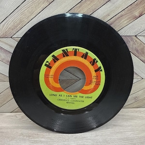 May include: A black vinyl record with a green, orange, and white label. The label reads "FANTASY" and "LONG AS I CAN SEE THE LIGHT" by Creedence Clearwater Revival. The record is a vintage music item.